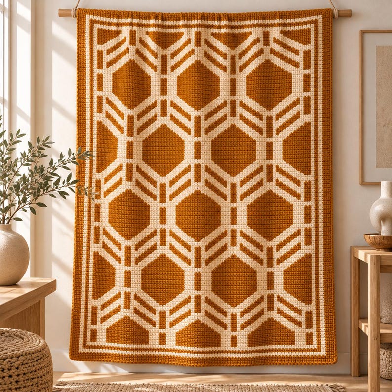 May include: A rectangular crochet wall hanging featuring a geometric pattern of interlocking hexagons in burnt orange and cream yarn. The textile is suspended from a wooden rod and displays a repeating symmetrical design with a thick border.