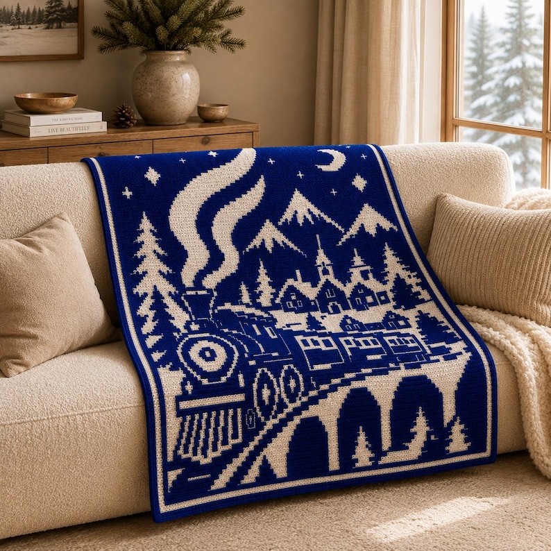 May include: A rectangular knit throw blanket featuring a deep blue background with a contrasting white graphic design. The pattern depicts a steam locomotive traveling across a bridge toward a village with mountains, pine trees, stars, and a crescent moon.