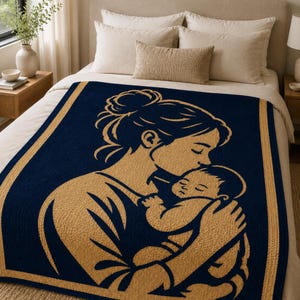 May include: Navy blue and tan blanket featuring a mother kissing a baby. The blanket has a gold border and a detailed illustration of a mother and child. The blanket is on a bed with white pillows and a beige comforter.