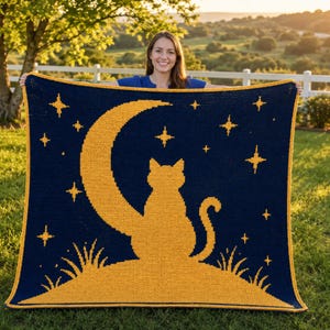 Op de afbeelding: Een marineblauwe gehaakte deken met een gele katten-silhouet op een maan en sterren. De deken heeft een gele rand. De deken wordt buiten gepresenteerd.