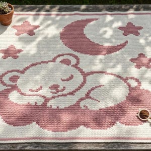 May include: A square crochet blanket featuring a cream-colored base with a pink graphic design of a sleeping bear resting on a cloud, surrounded by four stars and a crescent moon. The blanket has a simple pink border around the entire edge.