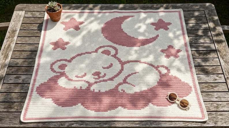 May include: A square crochet blanket featuring a cream-colored base with a pink graphic design of a sleeping bear resting on a cloud, surrounded by four stars and a crescent moon. The blanket has a simple pink border around the entire edge.