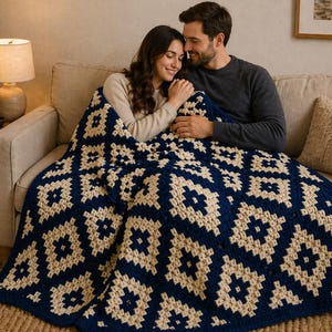 May include: A navy and cream-coloured crocheted blanket with a geometric pattern. The blanket is draped over a couple on a beige sofa. The blanket's design features repeating square motifs.