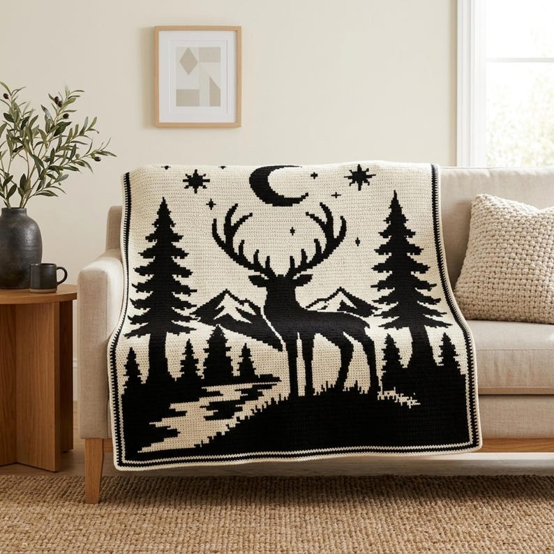 Peut inclure: Une couverture en crochet de couleur cr&egrave;me pr&eacute;sentant un motif graphique noir repr&eacute;sentant un cerf aux grands bois dans un paysage forestier avec des pins, des montagnes, un croissant de lune et des &eacute;toiles. La couverture a une bordure simple.