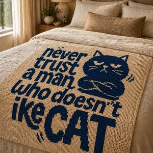 May include: Beige crocheted blanket with the text "Never trust a man who doesn't like CAT" in navy blue. A navy blue cat graphic is also featured on the blanket. The blanket is displayed on a bed.