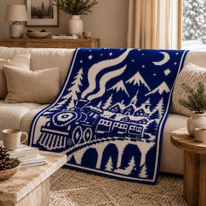 May include: A rectangular knit throw blanket featuring a deep blue background with a white, pixelated winter scene. The design depicts a steam locomotive pulling passenger cars across a bridge, with pine trees, mountains, stars, and a crescent moon.
