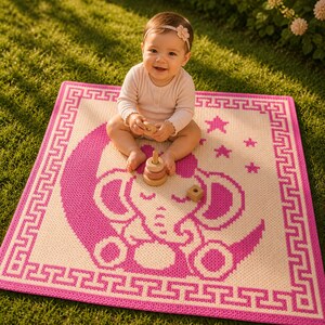 May include: A pink and cream-colored baby blanket with an elephant and moon design. The blanket has a Greek key border and star accents. A baby is sitting on the blanket with wooden toys.