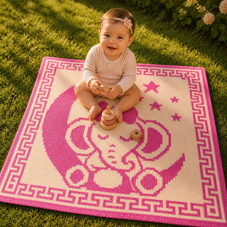 May include: A pink and cream-colored baby blanket with an elephant and moon design. The blanket has a Greek key border and star accents. A baby is sitting on the blanket with wooden toys.