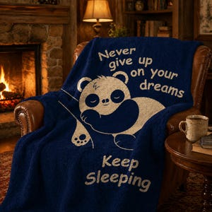May include: A navy blue throw blanket featuring a sleeping panda bear design and the text "Never give up on your dreams" and "Keep Sleeping" in white. The blanket is on a brown leather armchair.