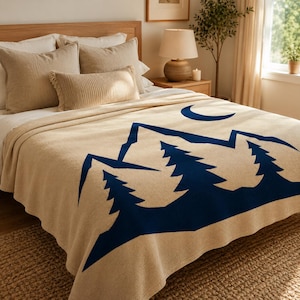 May include: A beige throw blanket featuring a dark blue graphic print of mountain peaks, pine trees, and a crescent moon. The blanket is draped over a bed with beige pillows, including two square pillows and one rectangular ribbed pillow.
