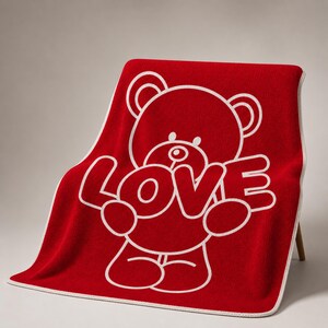 May include: A red blanket with a white outline of a teddy bear holding the word "LOVE". The blanket has a white border and is draped over a chair. The design is simple and bold.