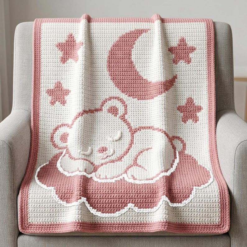 May include: A rectangular crochet blanket featuring a cream-colored base with a dusty rose border. The design displays a sleeping bear resting on a cloud, surrounded by a crescent moon and four stars, all rendered in a matching dusty rose yarn.