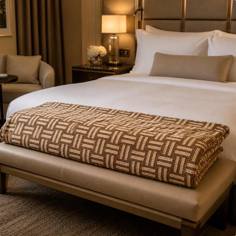 May include: A folded brown and beige woven blanket with a geometric pattern rests on a beige bench at the foot of a bed. The bed has white linens and a beige bolster pillow. A lamp and side table are visible.