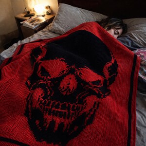 Puede incluir: Una manta de punto roja y negra con un gran dise&ntilde;o de calavera. La manta est&aacute; sobre una cama, con la cara de la calavera visible. La manta tiene un esquema de color rojo y negro, con un borde negro.