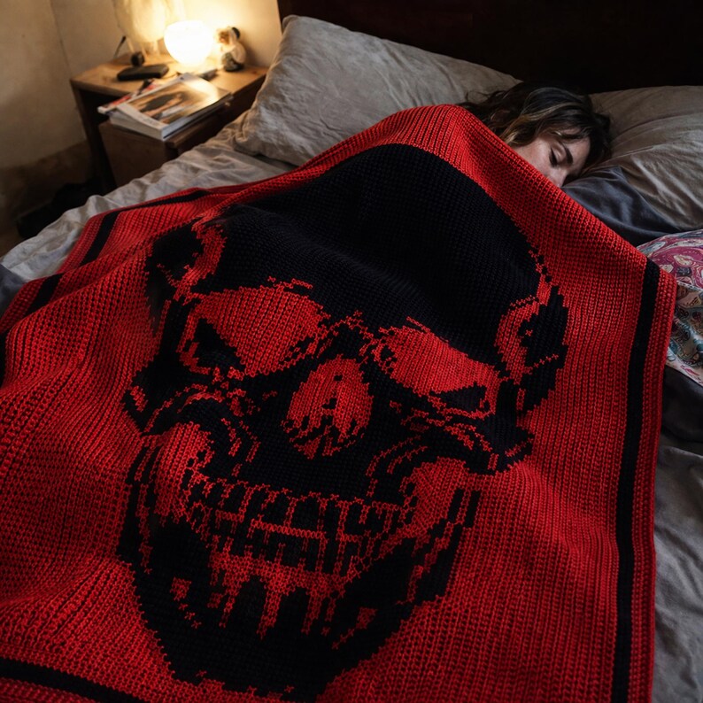 Puede incluir: Una manta de punto roja y negra con un gran dise&ntilde;o de calavera. La manta est&aacute; sobre una cama, con la cara de la calavera visible. La manta tiene un esquema de color rojo y negro, con un borde negro.