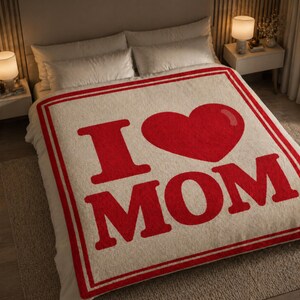 May include: A cream-colored blanket with a red border and the words "I ❤️ MOM" in large red letters. The blanket is on a bed with white sheets and pillows, with two lamps on either side.