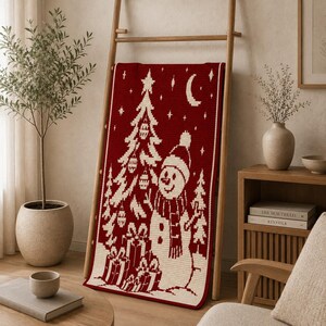 May include: A rectangular crochet blanket in deep red with a cream-colored pattern featuring a decorated Christmas tree, a snowman wearing a hat and scarf, stacked gift boxes, stars, and a crescent moon, displayed draped over a wooden ladder.
