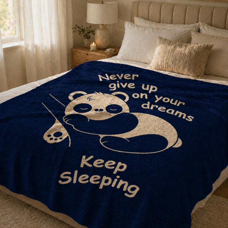 May include: A navy blue blanket featuring a sleeping panda bear illustration and the text "Never give up on your dreams" and "Keep Sleeping" in white. The blanket is on a bed with pillows.