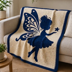May include: A cream-colored knitted blanket with a navy blue fairy silhouette holding a star. The blanket has a navy blue border and is draped over a beige sofa. The fairy has large wings and a flowing dress.