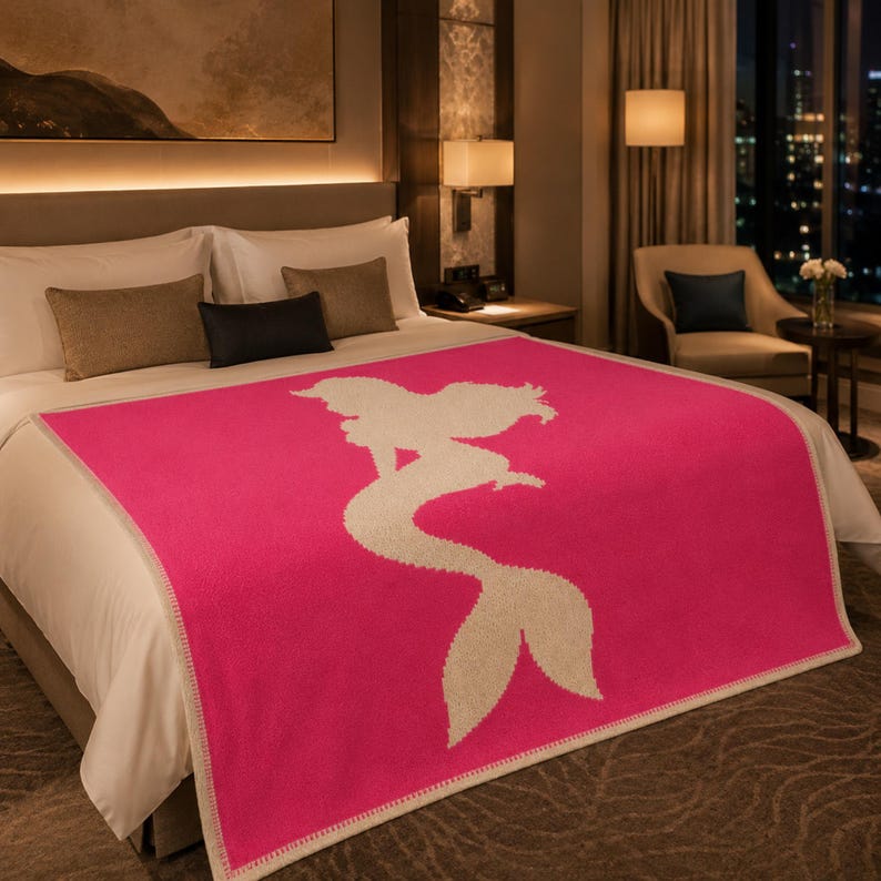 May include: A hot pink blanket with a cream-colored mermaid silhouette design. The blanket is on a bed with white sheets and brown pillows. The room has a warm, inviting atmosphere with soft lighting.