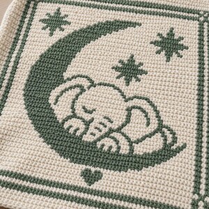 May include: A square crochet textile featuring a dark green design on a cream background. The pattern depicts a sleeping elephant resting on a crescent moon, surrounded by three eight-pointed stars and a small heart, all framed by a double border.