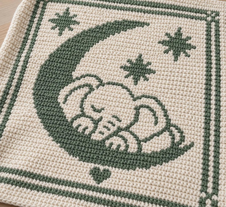May include: A square crochet textile featuring a dark green design on a cream background. The pattern depicts a sleeping elephant resting on a crescent moon, surrounded by three eight-pointed stars and a small heart, all framed by a double border.