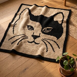 May include: A crocheted blanket featuring a cat face design. The blanket has a tan background with a black and white cat face. The border is black, and the cat's features are outlined in black. The blanket is square.