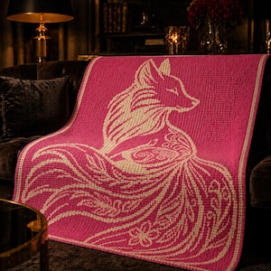 May include: A pink knitted throw blanket featuring a cream-colored fox design. The fox is depicted with intricate details, including a flowing tail and floral accents. The blanket is draped over a dark brown sofa, showcasing its texture and design.