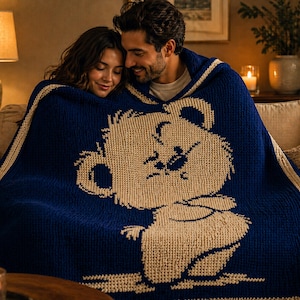 May include: A navy blue knitted blanket with a cream-coloured bear design. The blanket has a cream border and is draped over a couple. The blanket is made of a soft, textured material, ideal for snuggling on a sofa.