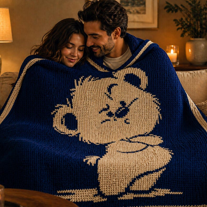 May include: A navy blue knitted blanket with a cream-coloured bear design. The blanket has a cream border and is draped over a couple. The blanket is made of a soft, textured material, ideal for snuggling on a sofa.