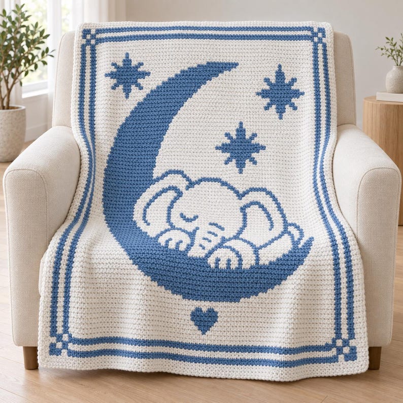 May include: A cream-colored crochet blanket featuring a blue design of a sleeping elephant resting on a crescent moon. The pattern includes three blue stars and a small blue heart at the bottom, all framed by a blue rectangular border.