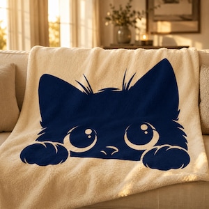 May include: A cream-colored sherpa blanket with a navy blue cat face design. The cat's large eyes and paws are visible, creating a playful and cozy aesthetic. The blanket is draped over a beige sofa.