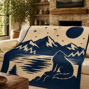 May include: A woven throw blanket featuring a dark blue and cream color scheme. The design depicts a wolf sitting by a lake, looking up at a large moon, with a mountain range, pine trees, and scattered stars in the night sky above the landscape.