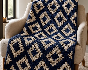 May include: A navy blue and cream-coloured crocheted blanket with a geometric diamond pattern draped over a beige armchair. The blanket's texture is visible, and the colour scheme creates a cosy, inviting aesthetic. The blanket is a home decor item.