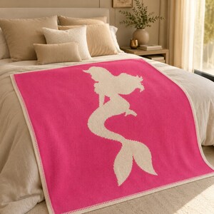 May include: A hot pink blanket featuring a white silhouette of a mermaid. The blanket has a white border and is displayed on a bed with neutral-toned pillows and bedding. The blanket is made of a soft material.