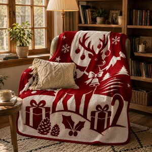 Pode incluir: Uma manta vermelha e branca com um grande gr&aacute;fico de rena com chifres, ornamentos e um sino. O design inclui caixas de presente, pinhas, folhas de azevinho e flocos de neve. Uma almofada de malha tran&ccedil;ada cor de creme repousa sobre ela.