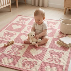 May include: A pink and cream-colored baby blanket with a teddy bear and heart pattern. A baby is sitting on the blanket, holding a stuffed teddy bear. A wooden rattle and a woven basket are nearby.