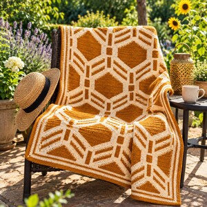 May include: A crochet blanket featuring a geometric pattern of mustard yellow and cream white hexagons. The blanket is draped over a dark chair next to a straw hat with a black band, a gold metal lantern, and a white ceramic mug.