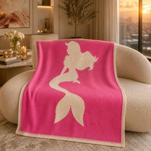 May include: A hot pink throw blanket with a cream-colored mermaid silhouette. The blanket is draped over a beige curved sofa. The blanket has a cream-colored border.