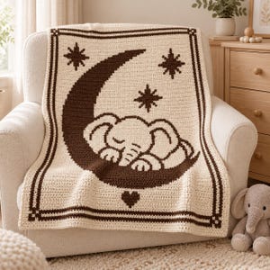 May include: A cream-colored crochet blanket featuring a dark brown border and a central graphic of a sleeping elephant resting on a large crescent moon. The design includes three eight-pointed stars and a small heart shape below the moon.