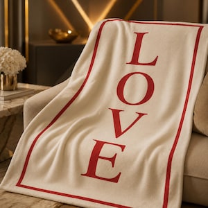 May include: Cream-colored blanket with the word "LOVE" in large red letters. The blanket has a red border and is draped over a beige sofa. The blanket appears soft and cozy, suitable for home decor.