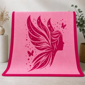 May include: A pink knitted blanket featuring a magenta fairy silhouette with large wings, butterflies, and stars. The blanket has a solid pink center with a magenta border. The design is a whimsical and decorative element.