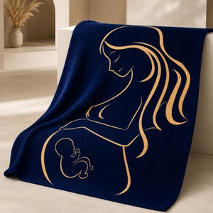 May include: A navy blue knitted blanket with a gold outline depicting a pregnant person and a baby. The blanket is folded over a white surface. The design is a soft, cozy item. The blanket is a soft, cozy item.