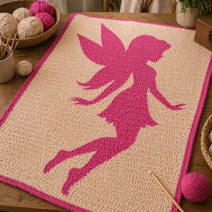 May include: A handmade crochet blanket featuring a pink fairy silhouette on a cream-colored background. The blanket has a pink border and is surrounded by yarn and crochet hooks. The blanket is approximately 30 inches by 40 inches.