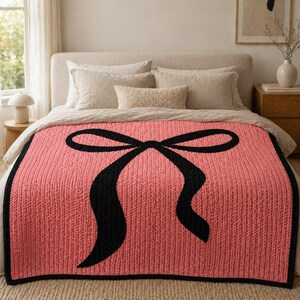 Pu&ograve; includere: Una coperta lavorata a maglia color corallo con un grande disegno a fiocco nero. La coperta ha una trama a coste e un bordo nero, che aggiunge un tocco di eleganza all'arredamento della camera da letto. La coperta &egrave; esposta su un letto con biancheria da letto dai toni neutri.