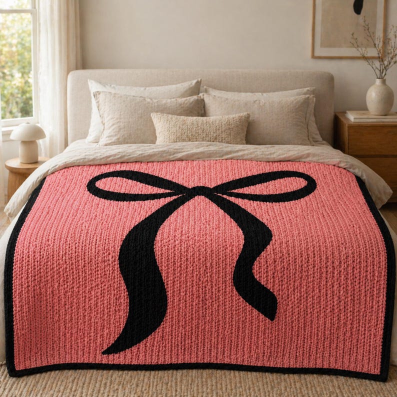 Pu&ograve; includere: Una coperta lavorata a maglia color corallo con un grande disegno a fiocco nero. La coperta ha una trama a coste e un bordo nero, che aggiunge un tocco di eleganza all'arredamento della camera da letto. La coperta &egrave; esposta su un letto con biancheria da letto dai toni neutri.