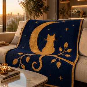 May include: A navy blue crochet blanket featuring a golden cat silhouette perched on a crescent moon and branch, with stars. The blanket is draped over a beige sofa, creating a cozy, celestial-themed home decor item.