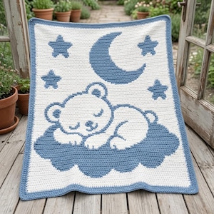May include: A rectangular crochet blanket featuring a white background with a blue border. The design displays a white bear sleeping on a blue cloud, surrounded by a blue crescent moon and four blue stars, all crafted with a consistent stitch pattern.