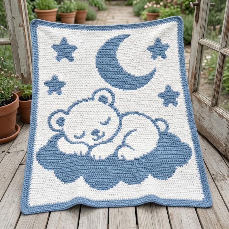 May include: A rectangular crochet blanket featuring a white background with a blue border. The design displays a white bear sleeping on a blue cloud, surrounded by a blue crescent moon and four blue stars, all crafted with a consistent stitch pattern.