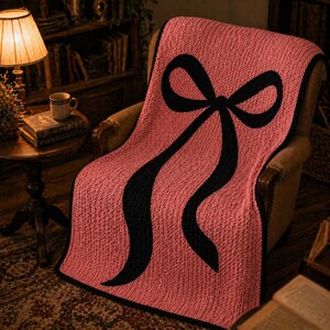 Pu&ograve; includere: Una coperta rosa lavorata a maglia con un grande disegno a fiocco nero, drappeggiata su una sedia. La coperta ha un aspetto strutturato e un bordo nero. I nastri del fiocco scendono, creando un contrasto sorprendente.